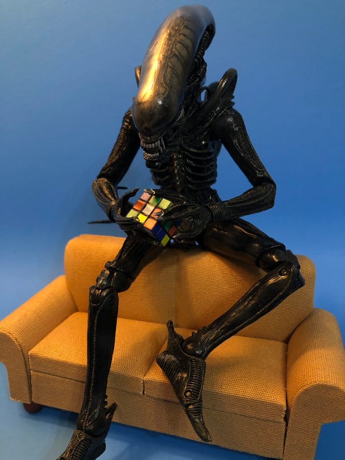 A Xenomorph sits on a couch playing with a Rubik's cube.