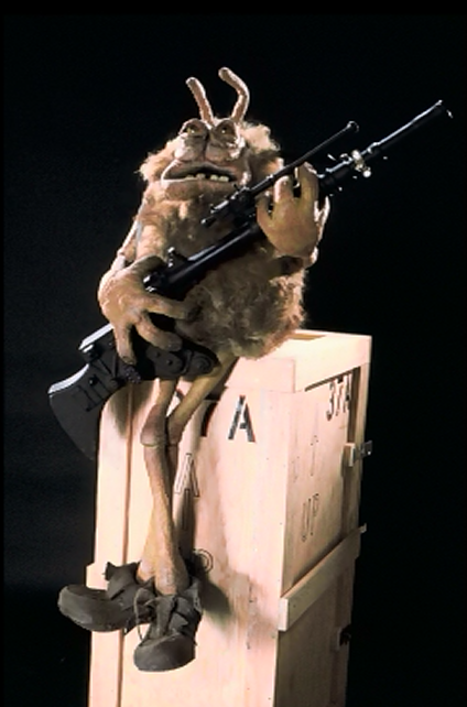 A weird Muppet-like alien holds a rife while sitting on a crate.