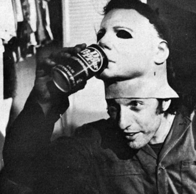 A guy tips a Dr Pepper towards the Michael Myers mask that's pulled up on his own forehead, as if giving it a drink.