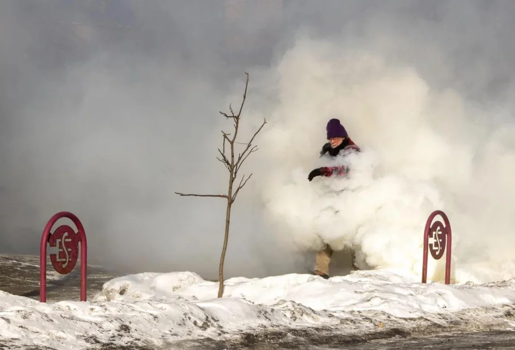 A bearded old guy with glasses pushes through a cloud of white smoke, walking along a snow-covered sidewalk near a tiny bare tree.